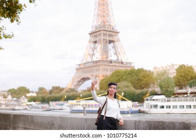 Smiling African American Taking Selfies On Background Of Eiffel Tower Using Silver Smartphone. Young Woman Came To Paris For Honeymoon. Black-haired Woman With Beautiful Hair Styling Wearing