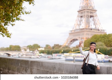 Smiling African American Taking Selfies On Background Of Eiffel Tower Using Silver Smartphone. Young Woman Came To Paris For Honeymoon. Black-haired Woman With Beautiful Hair Styling Wearing