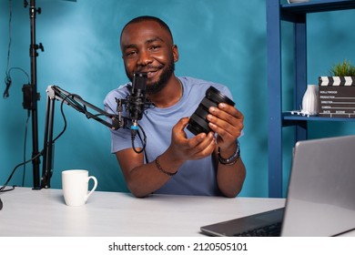 Smiling african american techology reviewer presenting dslr zoom lens features in live online podcast. Famous influencer with microphone reviewing photography equipment in front of laptop.