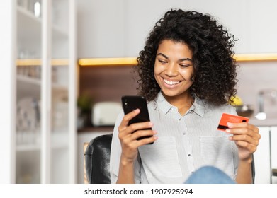 Smiling African American teenage girl with curly hair holding mobile phone, entering credit card number to make an online transaction, mixed-race woman ordering food, doing online shopping from home