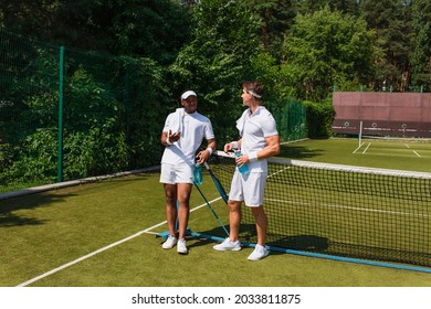 Smiling african american tennis player talking to friend with bottle of water on court