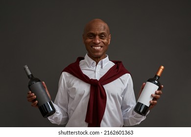 Smiling african american winemaker or sommelier with two wine bottles looking at camera. Adult successful male entrepreneur. Viticulture and winemaking. Grey background. Studio shoot. Copy space