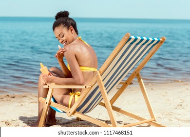 smiling african american woman applying sunscreen lotion on skin while sitting on deck chair on sandy beach  - Powered by Shutterstock