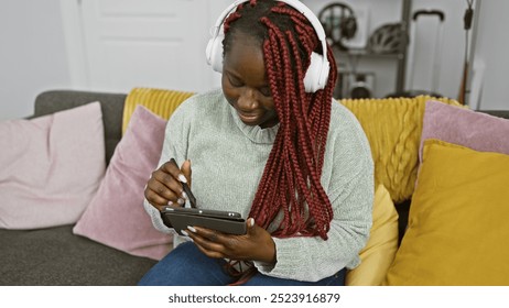 Smiling african american woman with braids wearing headphones uses smartphone at home on couch with colorful pillows. - Powered by Shutterstock