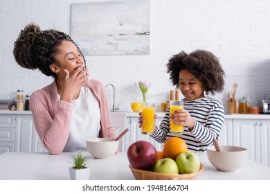 smiling african american woman covering mouth while daughter holding orange juice in kitchen, blurred foreground - Powered by Shutterstock