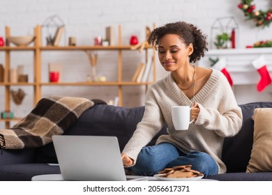 Smiling African American Woman With Cup Using Laptop Near Christmas Cookies