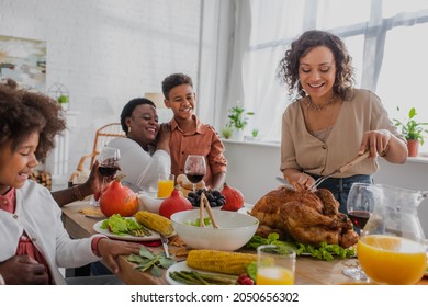 Smiling African American Woman Cutting Turkey Near Family And Thanksgiving Dinner