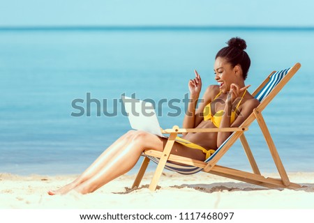 smiling african american woman gesturing by fingers and communicating by laptop while sitting on deck chair on sandy beach 