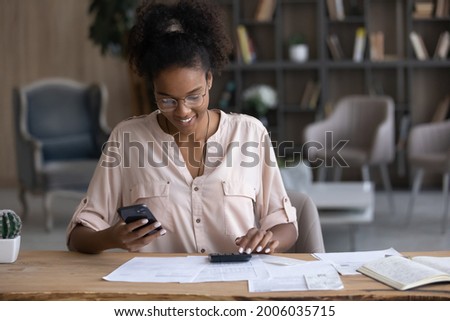 Smiling African American woman in glasses calculating bills, using smartphone and calculator, positive young female browsing online banking service, analyzing financial documents, planning budget