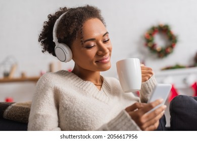 Smiling african american woman in headphones holding cup and cellphone during christmas at home - Powered by Shutterstock