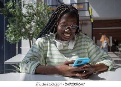 Smiling African American woman with headphones using her smartphone, enjoying sunny day and staying connected. Black girl scroll through news feed on her phone while sitting at table at mall - Powered by Shutterstock