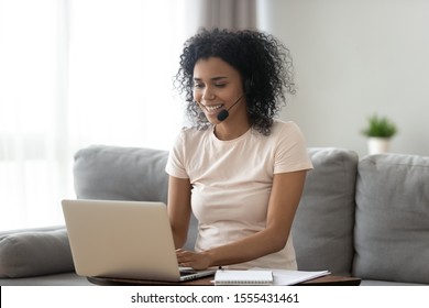 Smiling African American Woman In Headset Using Laptop, Talking, Working Customer Support Service Operator At Home, Girl In Headphones With Microphone Sitting On Couch, Studying Online