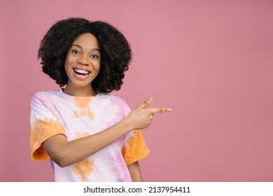 Smiling African American Woman  Looking At Camera Pointing Finger On Copy Space. Happy Curly Haired Fashion Model Wearing Stylish Tie Dye T Shirt Isolated On Pink Background. Shopping, Sale Concept 