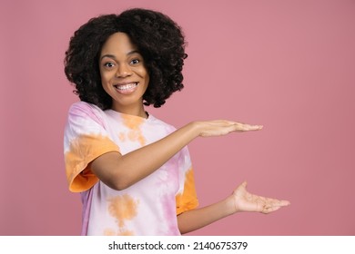 Smiling African American Woman Looking At Camera Holding Hands Near Copy Space. Happy Curly Haired Fashion Model Wearing Stylish Tie Dye T Shirt Isolated On Pink Background. Shopping, Sale Concept