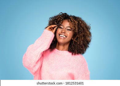 Smiling African American Woman In Pink Sweater And Eyeglasses Posing On Blue Studio Background .