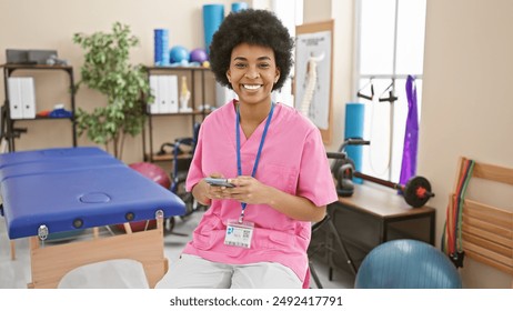 A smiling african american woman in pink scrubs using a smartphone in a well-equipped physical therapy clinic. - Powered by Shutterstock