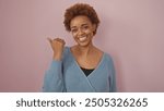 Smiling african american woman pointing sideways in a cozy sweater against a pink background.