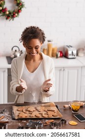 Smiling African American Woman Pouring Flour On Christmas Cookies Near Smartphone And Tea