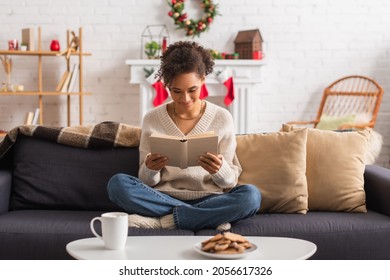 Smiling African American Woman Reading Book Near Christmas Cookies And Cup At Home