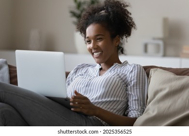 Smiling African American woman relax on sofa talk speak on video call on lapto. Happy young ethnic female havep fun laugh engaged in webcam digital virtual event on computer. Technology concept.