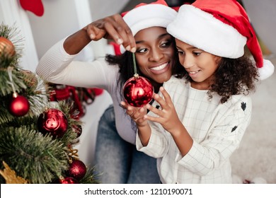 smiling african american woman in santa claus hat helping daughter to decorate christmas tree at home