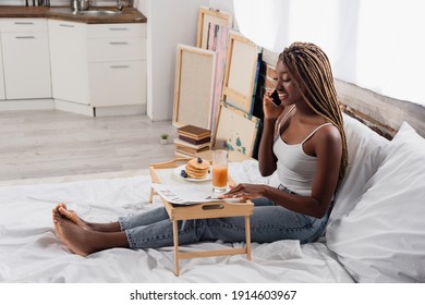 Smiling african american woman talking on smartphone near newspaper and breakfast on tray on bed