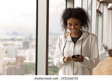 Smiling African American woman using phone, standing near panoramic window, happy young female looking at phone screen, browsing apps, surfing internet, chatting or shopping online, having fun