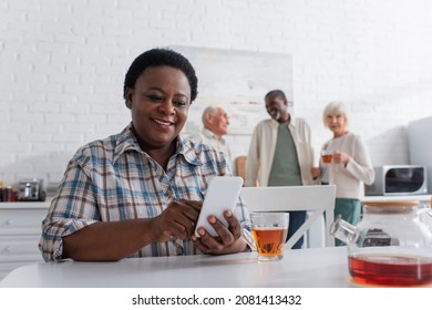 Smiling african american woman using smartphone near tea and blurred people in nursing home - Powered by Shutterstock