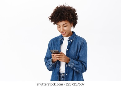 Smiling african american woman using mobile phone, holding telephone and looking happy, standing over white background - Shutterstock ID 2237217507