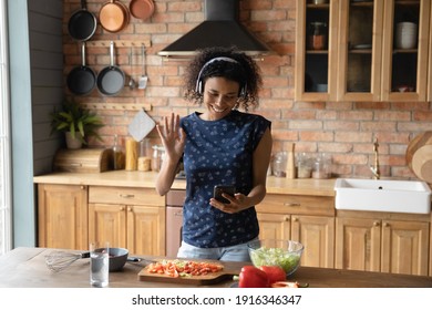 Smiling African American woman wearing headphones making video call, standing in kitchen, chatting with friends or relatives online, waving hand, blogger shooting vlog, cooking, preparing salad