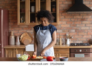 Smiling African American woman wearing apron cutting fresh vegetables on wooden baord, cooking salad, standing in kitchen at home, happy young female preparing dinner, organic vegetarian snack