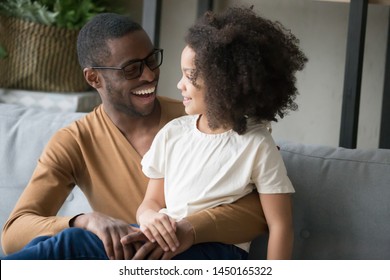 Smiling African American Young Dad Sit On Couch Hug Cute Little Daughter Chatting Or Talking, Happy Black Father Spend Time Having Conversation With Funny Preschooler Child, Enjoy Weekend Together