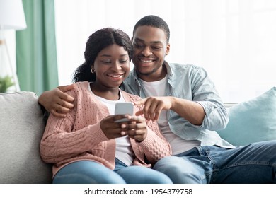 Smiling African American Young Family Using Mobile Phone Together While Resting On Couch In Living Room, Copy Space. Happy Black Couple Checking New Shopping Application On Smartphone