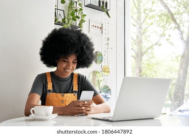 Smiling African American young hipster mixed race gen z young woman with Afro hair holding smart phone texting, using mobile app watching video sitting at table in cozy modern cafe with laptop. - Shutterstock ID 1806447892