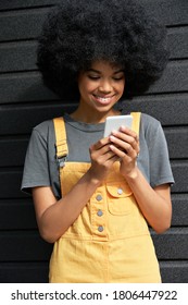 Smiling African American Young Hipster Lady With Afro Hair Standing On Black Background Holding Smart Phone. Happy Young Mixed Race Gen Z Woman Using Mobile Social Network Media App, Vertical.