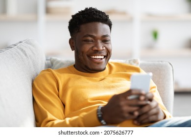 Smiling african american young man reclining on couch with cellphone, chatting with ladies, using dating mobile application on cellphone, home interior, closeup portrait, copy space - Powered by Shutterstock