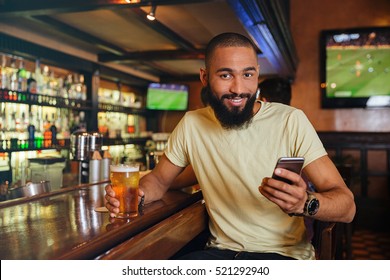 Smiling African American Young Man Drinking Beer And Using Cell Phone In Pub