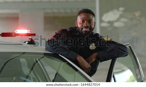 Smiling african american young man cops stand near\
patrol car look at camera enforcement happy officer police uniform\
auto safety security communication control policeman close up slow\
motion