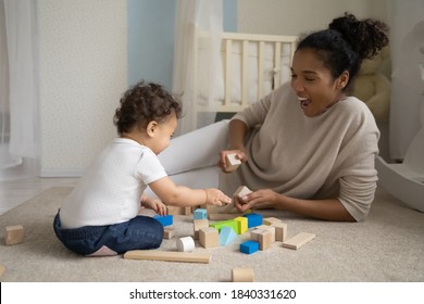 Smiling african American young mom lying on floor at home kids room play with building blocks with little baby toddler. Happy loving caring biracial mother engaged in funny activity with infant child.