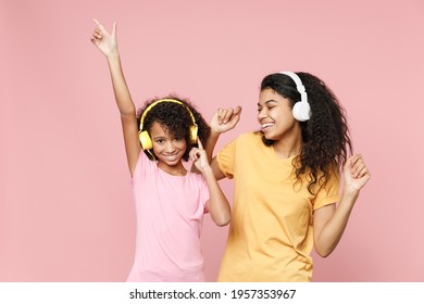 Smiling african american young woman and little kid girl sisters in basic t-shirts listening music with headphones dancing rising hands isolated on pink background studio portrait. Family day concept