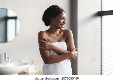 Smiling African American Young Woman Covered In White Towel Posing At Beautiful Bathroom, Hugging Her Shoulders And Looking At Copy Space, Enjoying Her Silky Skin. Body, Skin Care Concept