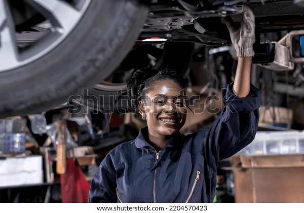 Smiling African black mechanic woman in overall\
clothing happy working underneath lifted vehicle in car repair\
service shop, black auto technician woman fixing vehicle,car\
maintenance repair\
concept