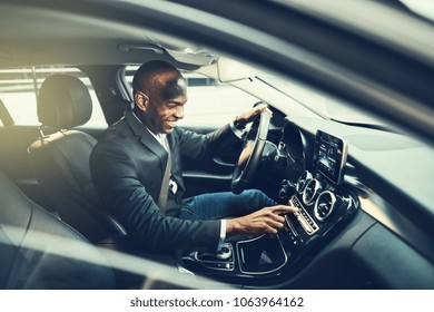 Smiling African Businessman Changing Stations On His Car Radio While Driving Through The City During His Morning Commute To Work