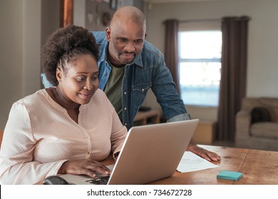 Smiling African Couple Doing Their Online Banking And Paying Bills With A Laptop At Their Kitchen Table At Home