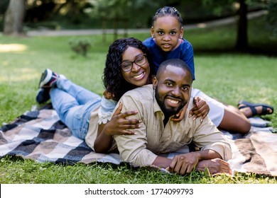 Smiling African Family And Small Child Girl Having Fun Together Outdoors In Spring Or Summer Nature In Park, Resting On Checkered Blanket On Green Meadow. Happy Family Concept