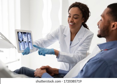 Smiling African Female Dentist Showing Young Man Patient Teeth Xray, Using Digital Tablet