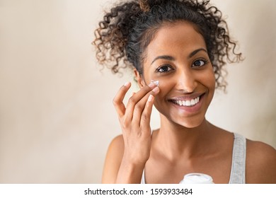 Smiling african girl with applying facial moisturizer while holding jar and looking at camera. Portrait of young black woman applying cream on her face isolated on beige background with copy space.