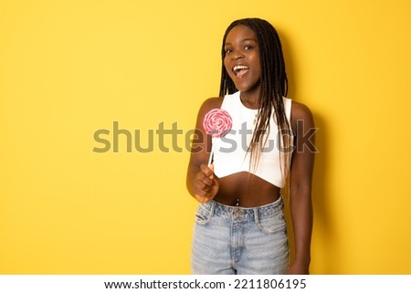 Smiling african girl holding candy lollipop. Young cheerful woman with lollipop isolated over yellow background,