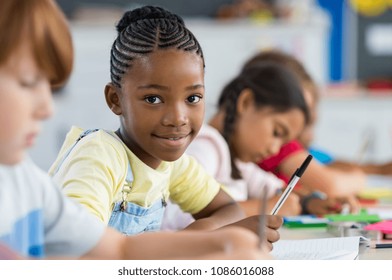 Smiling african girl sitting at desk in class room and looking at camera. Portrait of young black schoolgirl studying with classmates in background. Happy smiling pupil writing on notebook. 