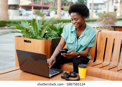 Smiling African girl sitting in a street cafe and working at the computer. The girl checks the data on the laptop and in the phone. Work online, business online